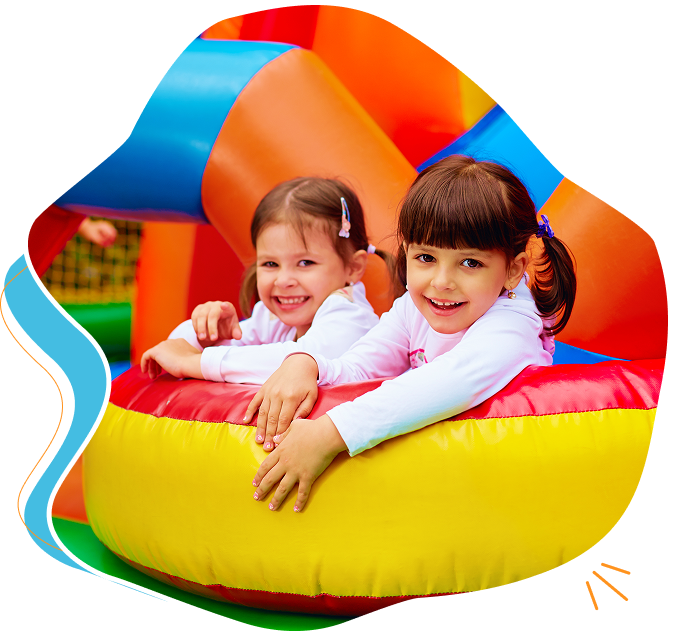 Two young girls smile while leaning on a colorful inflatable structure, enjoying some Bouncy House Fun at an indoor play area.